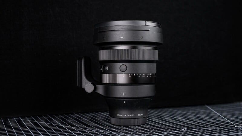 A black Sigma camera lens with a tripod mount is standing upright on a grid-patterned surface against a plain black background.