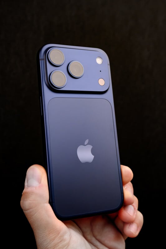 A hand holding a blue Apple iPhone with three rear cameras, a flash, and the Apple logo visible on the back against a dark background.