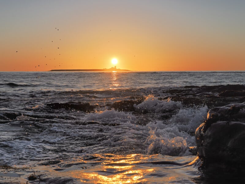 Waves crashing on rocky shore at sunset, with the sun low on the horizon above a distant island, casting golden light across the water and sky.