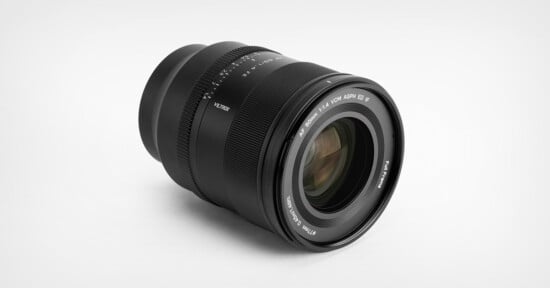 A black Sigma camera lens with white text markings, placed on a plain white surface, positioned at an angle to show the glass element and focus ring.