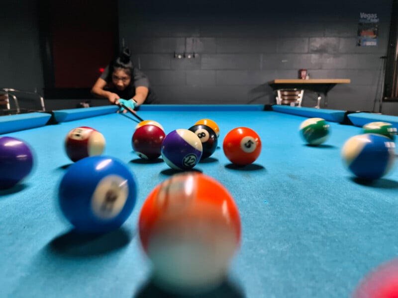 A person aims a cue stick at a blue pool table, striking the cue ball. Several colorful billiard balls scatter across the table in motion, with the background slightly out of focus.