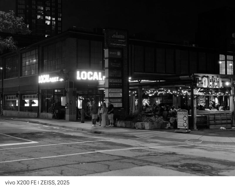 A black and white night photo of a corner restaurant called LOCAL, with illuminated signs, people gathered outside, and string lights on the patio. The street in front is empty and quiet.