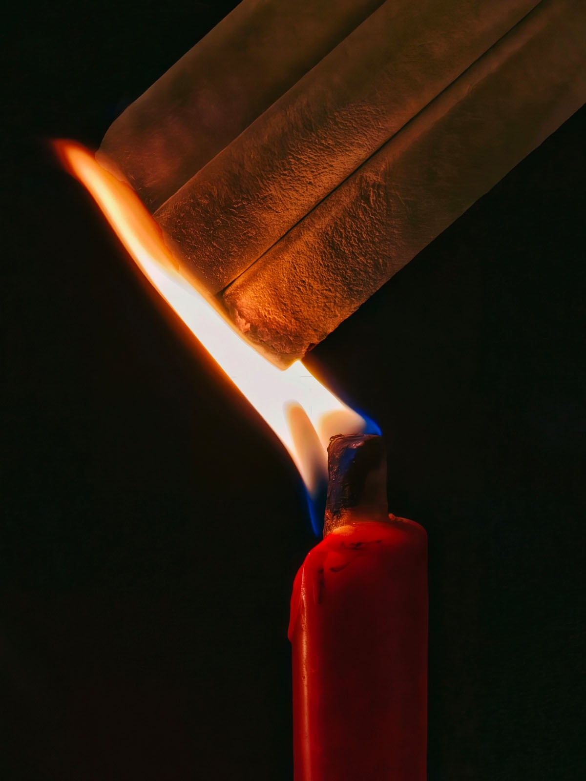 A close-up of a lit red candle with a bright flame melting the end of a translucent stick, set against a dark background.