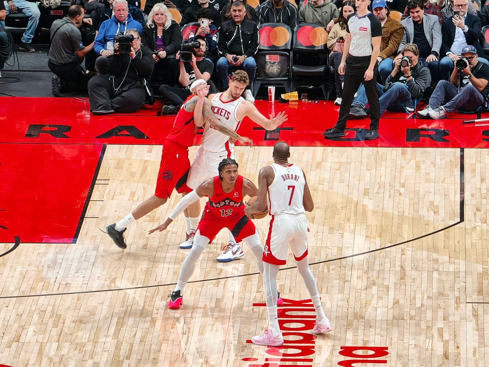 Two basketball players in red uniforms defend against a player in a white uniform holding the ball, while photographers and spectators watch from the sidelines of a brightly lit indoor court.