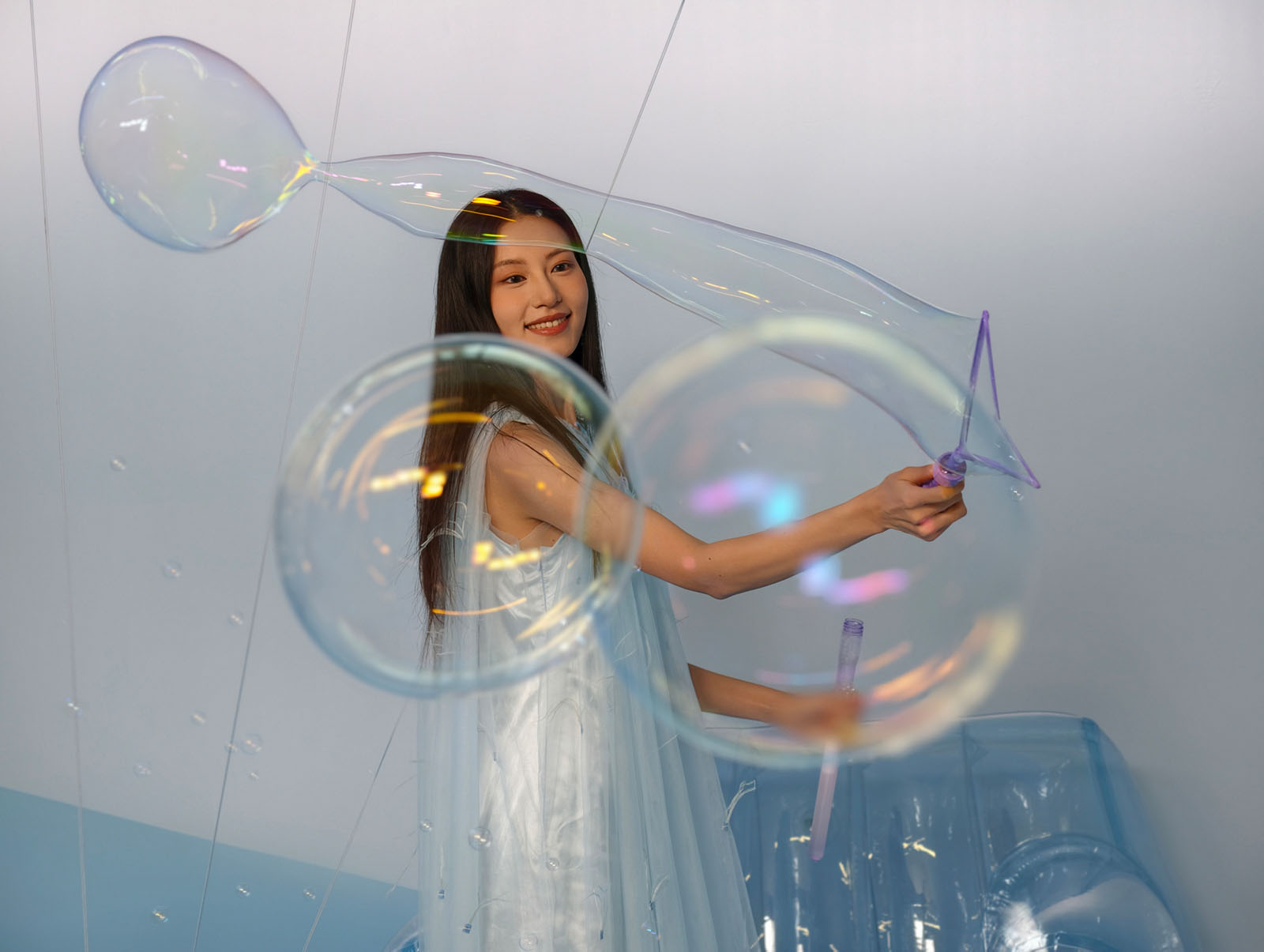 A young woman in a light, flowing dress smiles while creating large soap bubbles with a purple wand, surrounded by several floating bubbles against a soft, light blue and white background.