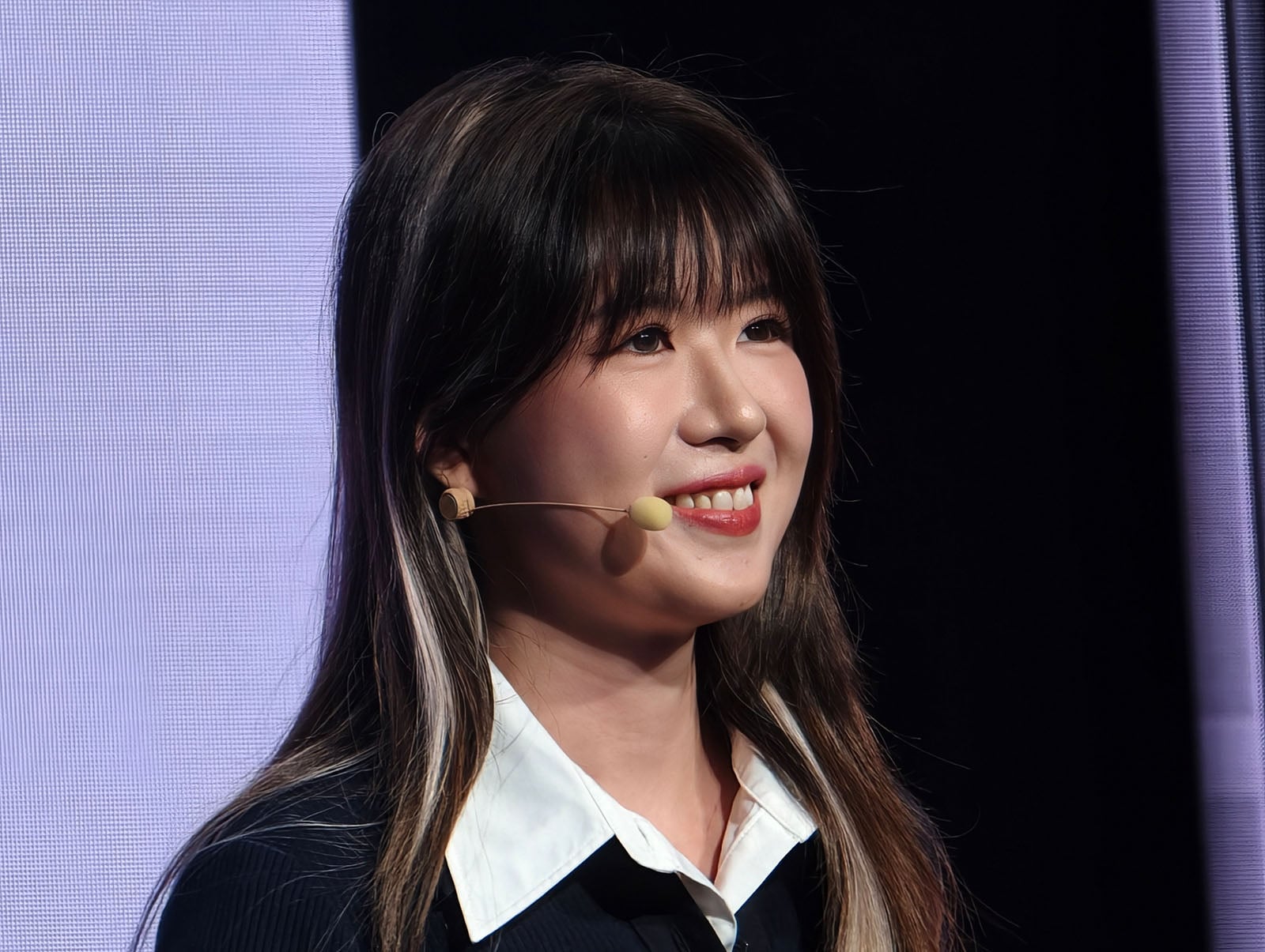 A woman with long brown hair and bangs, wearing a black sweater over a white collared shirt and a headset microphone, is smiling while standing in front of a lit screen and dark background.