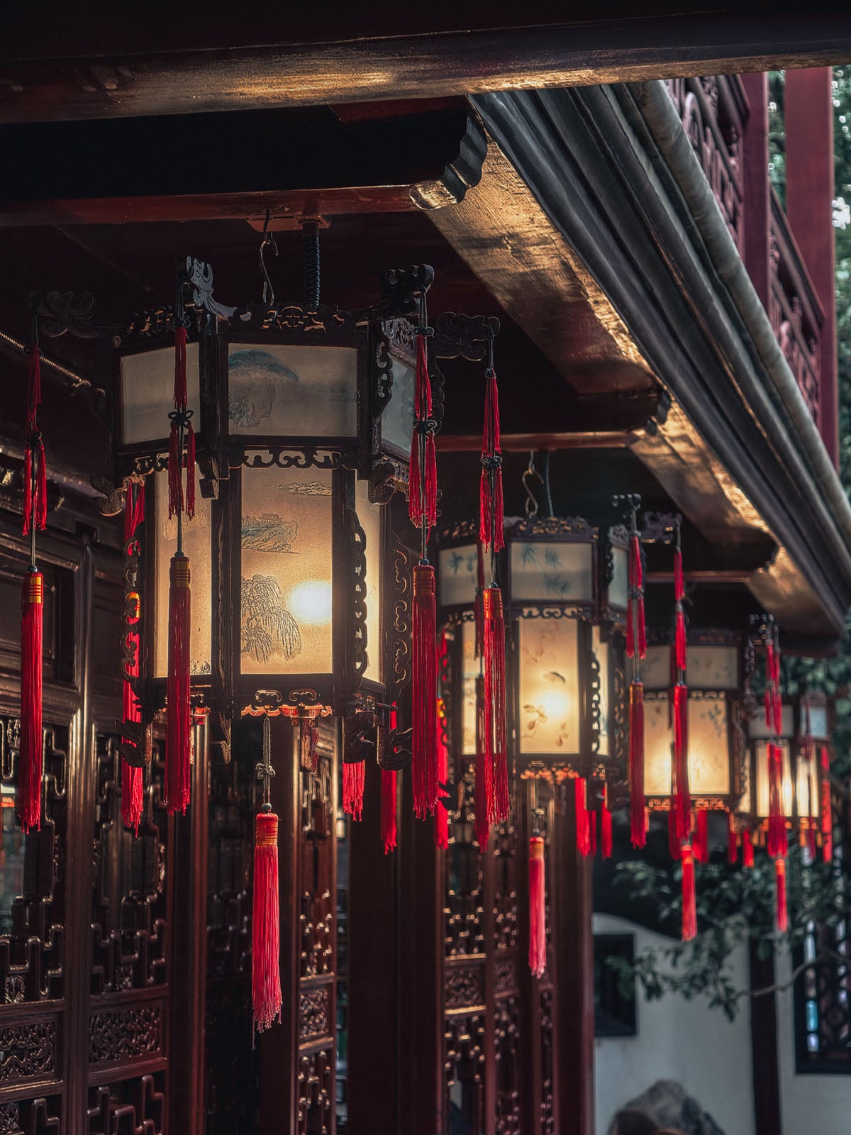 Traditional Chinese lanterns with ornate designs and red tassels hang in a row under a wooden roof, softly illuminated and casting a warm glow along a wooden building facade.