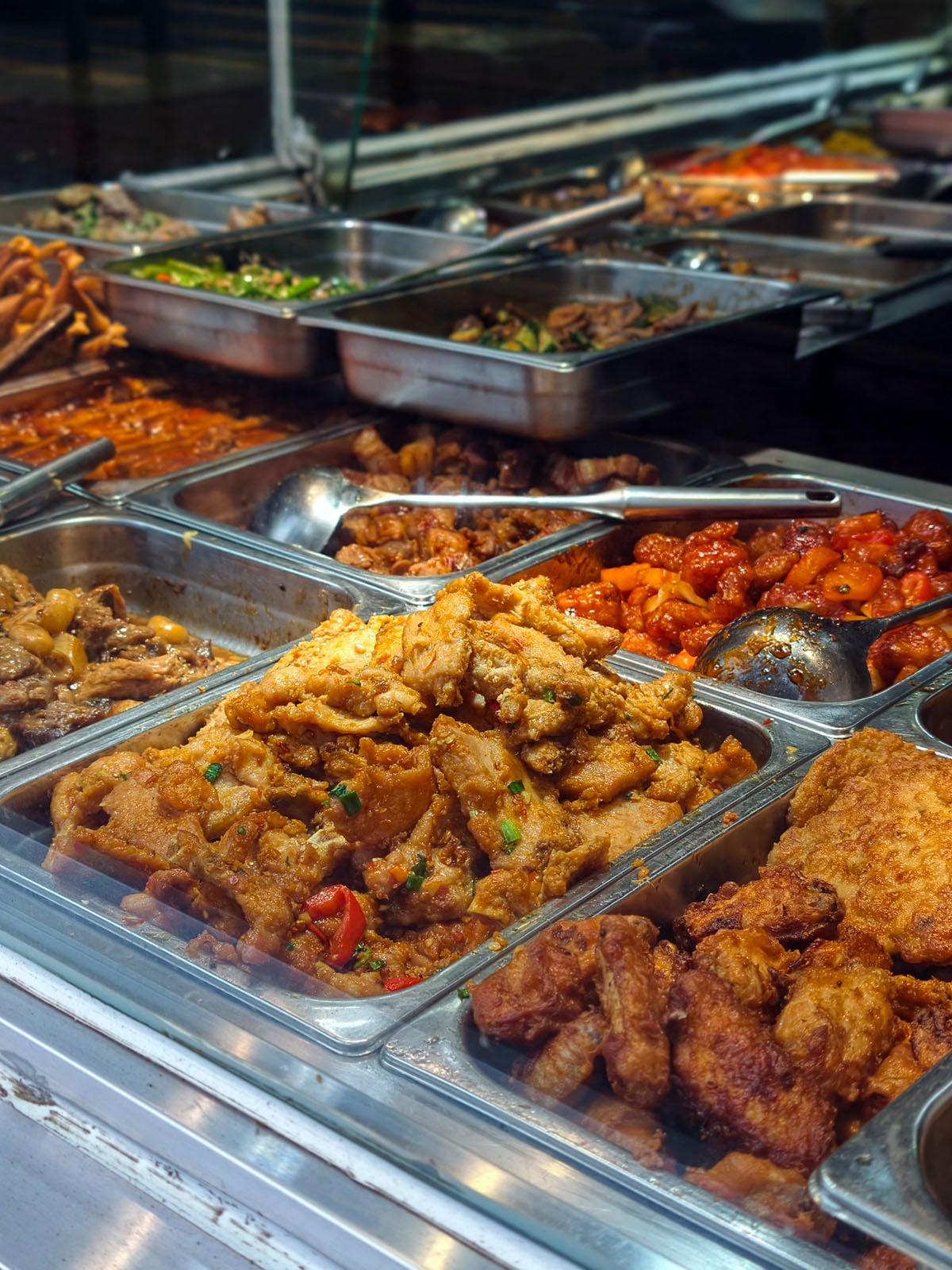 An assortment of cooked dishes in metal trays at a food stall, featuring fried foods, vegetables, and various stir-fried meats, all displayed behind a glass counter.