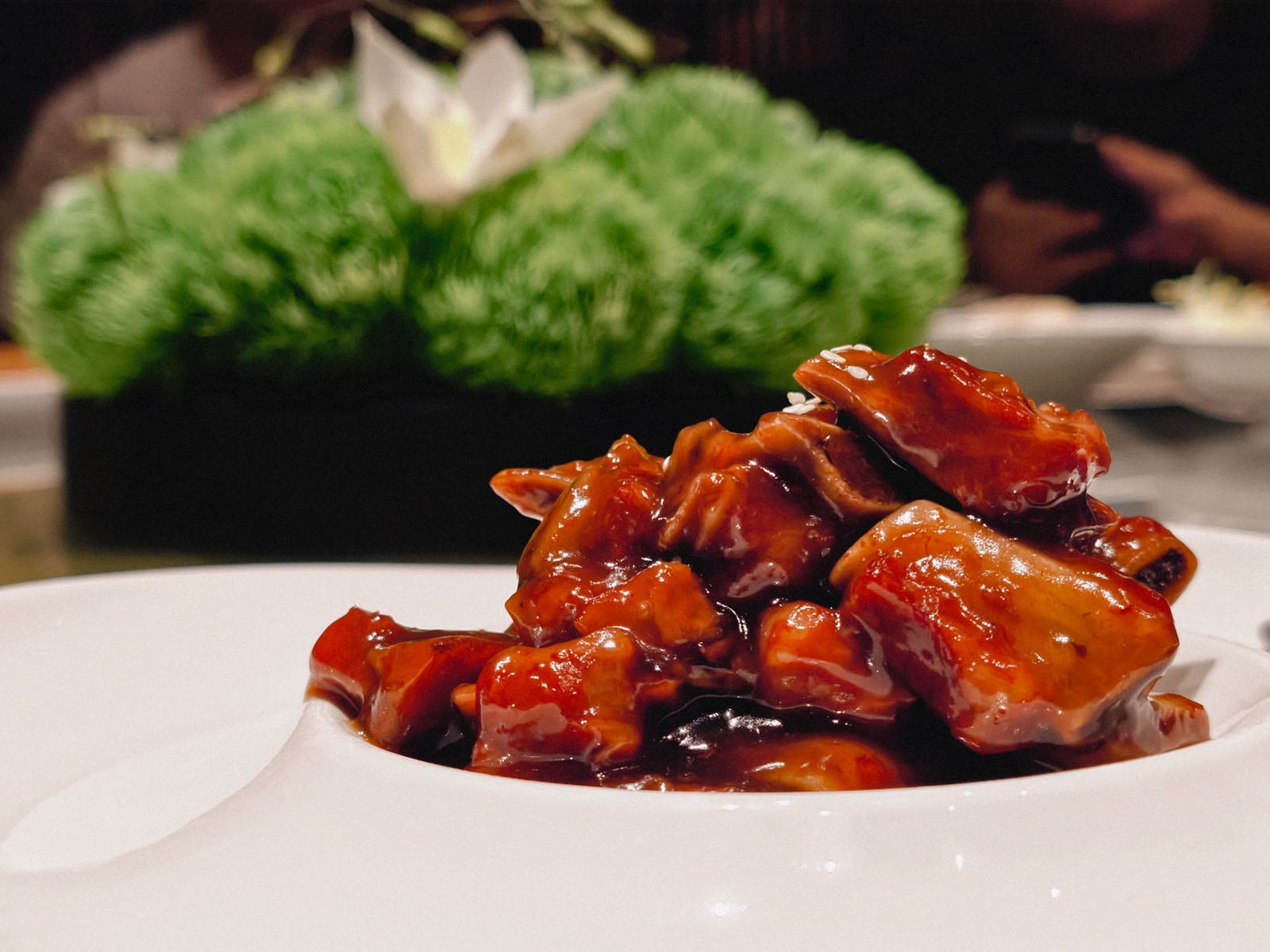 A close-up of a plate of glazed meat in a rich, dark sauce, with a blurred green floral centerpiece in the background. The food is presented on a white dish.