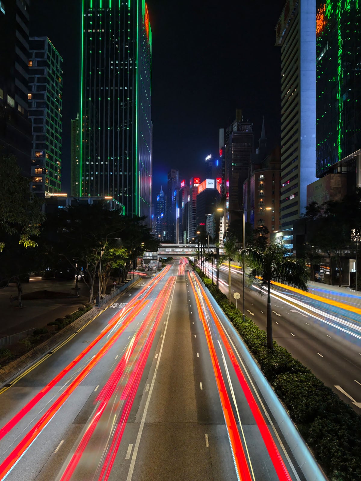A city street at night with tall buildings lit by colorful neon lights. Long-exposure photo shows red and white light trails from moving vehicles, emphasizing the urban energy and motion.