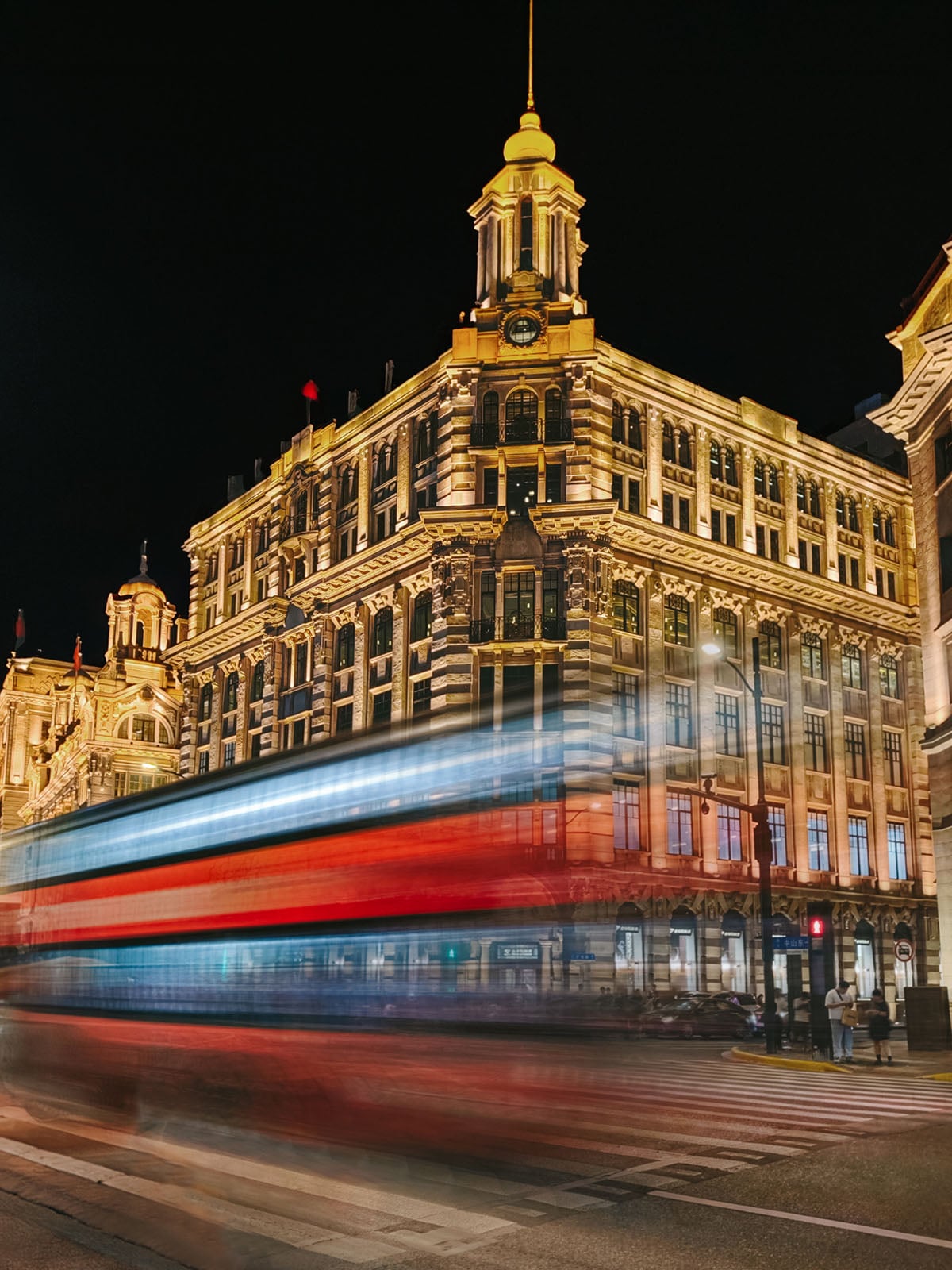 A historic, ornate building is illuminated at night as a bus, captured with a long exposure, creates colorful streaks of red, white, and blue light in front of it on a city street.