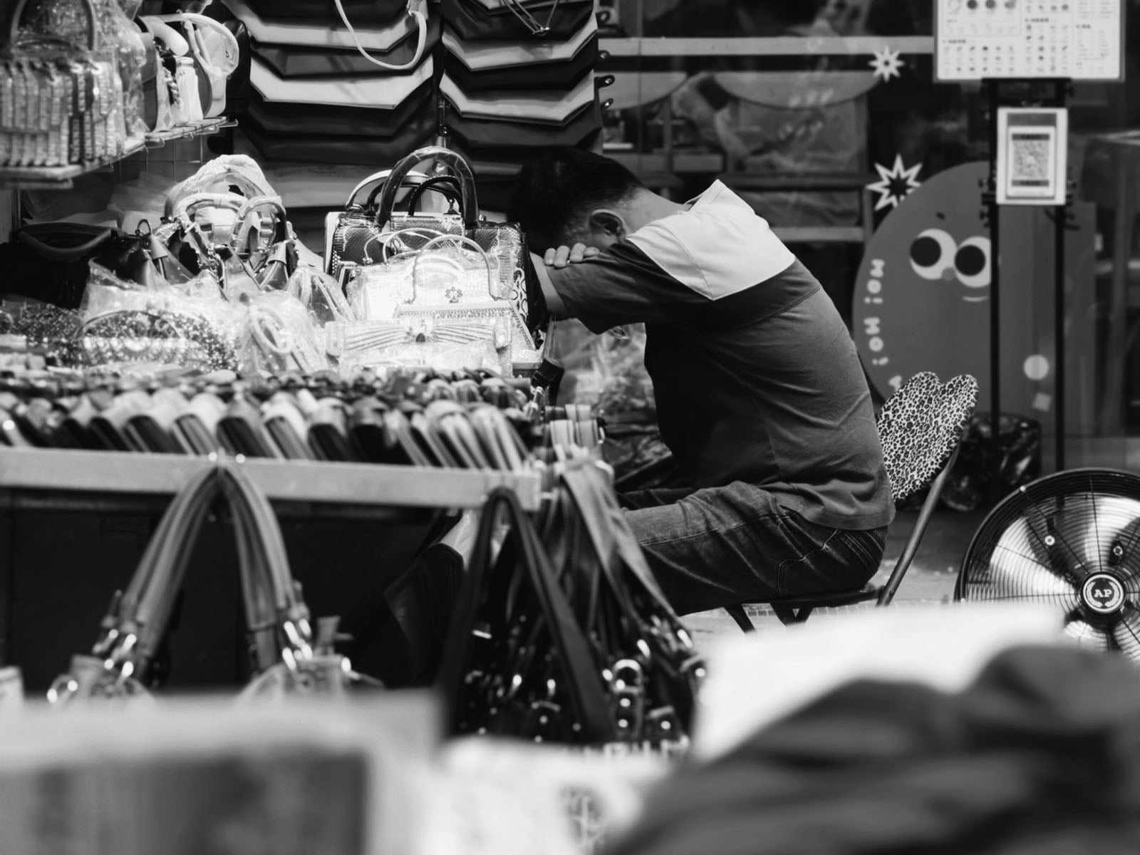 A man sits at a market stall with his head resting on his arms, appearing to nap. Handbags and purses are displayed on tables around him. The scene is in black and white.