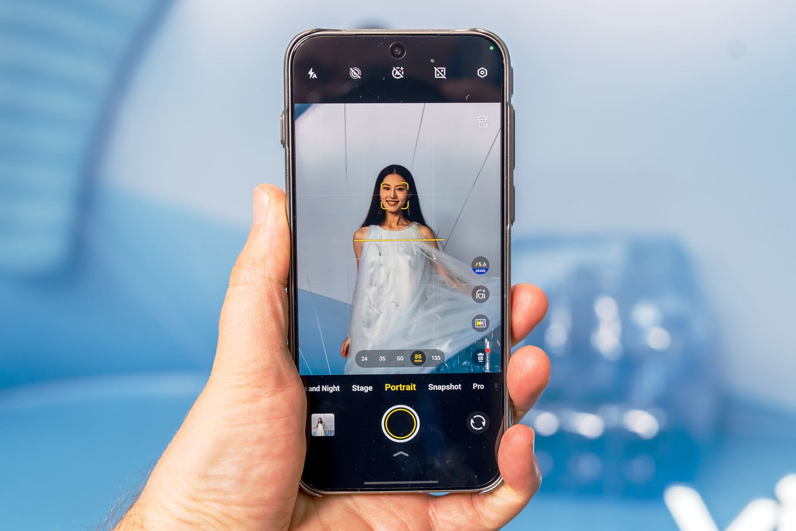 A hand holds a smartphone displaying its camera app, focused on a woman in a white dress posing against a blue background. The phone screen shows photo options and portrait mode settings.