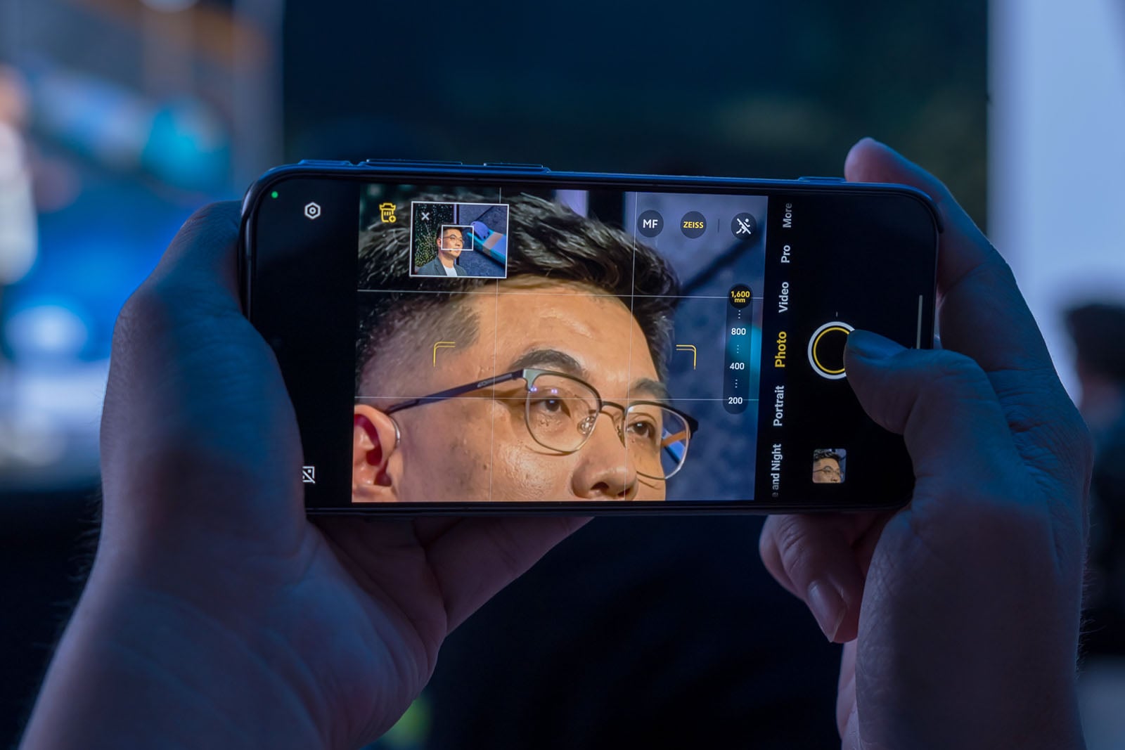Close-up of hands holding a smartphone, taking a photo of a man's face. The phone screen shows the camera app focused on the man's face with glasses, capturing his portrait in a well-lit indoor setting.