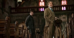 Two men stand in the aisle of an old, dimly lit church with wooden pews and stained glass windows; one wears a brown suit holding a hat, the other wears dark clothes and looks towards him.