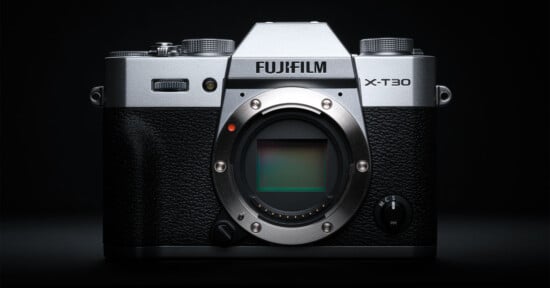 A Fujifilm X-T30 mirrorless digital camera with a silver and black body is shown without a lens, revealing the camera sensor, against a dark background.
