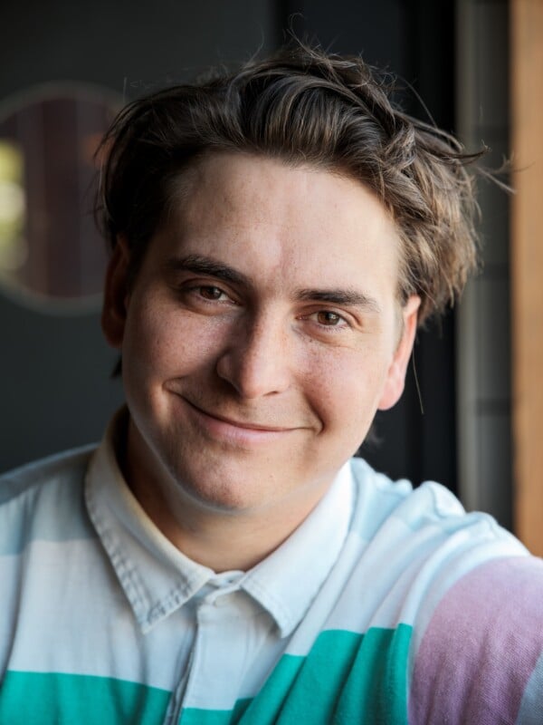 A person with short, tousled brown hair smiles warmly at the camera. They are wearing a white collared shirt with teal and purple stripes, and are sitting indoors near a window.