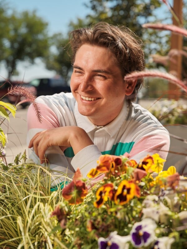 A person with brown hair and a striped shirt smiles while sitting outdoors next to colorful flowers and greenery on a sunny day.
