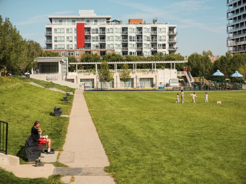 A grassy park with a walking path, benches, and a playground. Two people sit on a bench in the foreground, while several children and a dog play on the field. Apartment buildings are visible in the background.