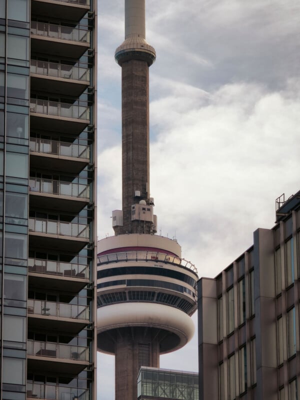 The CN Tower in Toronto rises between two modern high-rise buildings, with its observation deck and upper structure visible against a cloudy sky.