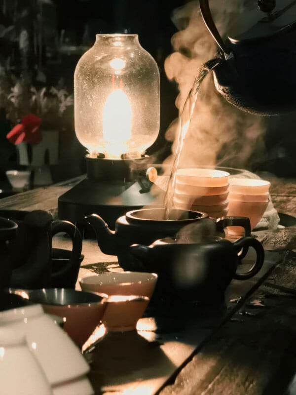 A warm lantern lights a rustic wooden table set for tea, with steam rising as hot water is poured from a kettle into stacked cups, surrounded by teapots and bowls.