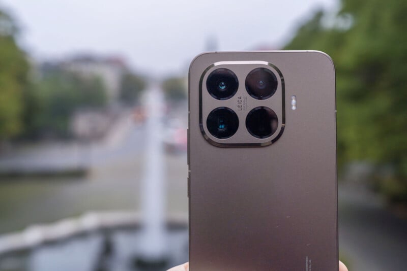 A close-up of a smartphone's rear cameras, featuring four lenses in a square arrangement. The phone is held outdoors with a blurred background of trees and a street.