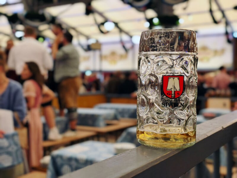 A large glass beer stein with the Spaten logo sits on a railing, nearly empty, in a lively beer hall with people and tables in the background.