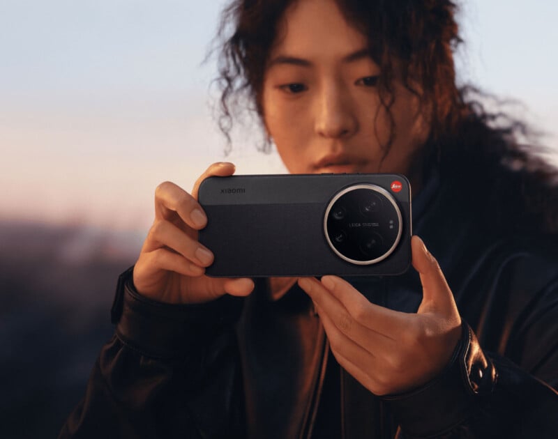 A person with curly hair and a leather jacket is holding a Xiaomi smartphone with a large circular camera module, focusing intently as they prepare to take a photo outdoors at sunset.