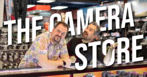 Two men stand behind a counter in a camera store, smiling at the camera. Large white text across the image reads "THE CAMERA STORE." Cameras and accessories are visible in the background.