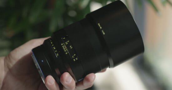 A hand holding a black camera lens with yellow markings and numbers, against a blurred background with green leaves and natural light.