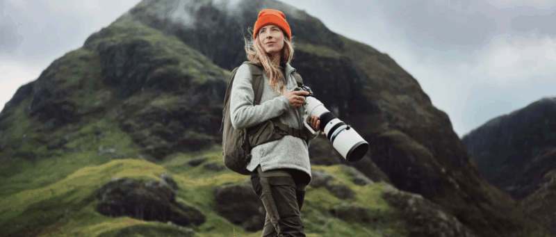 A woman wearing an orange beanie and gray jacket stands outdoors holding a camera with a large lens, with a green mountain and cloudy sky in the background.